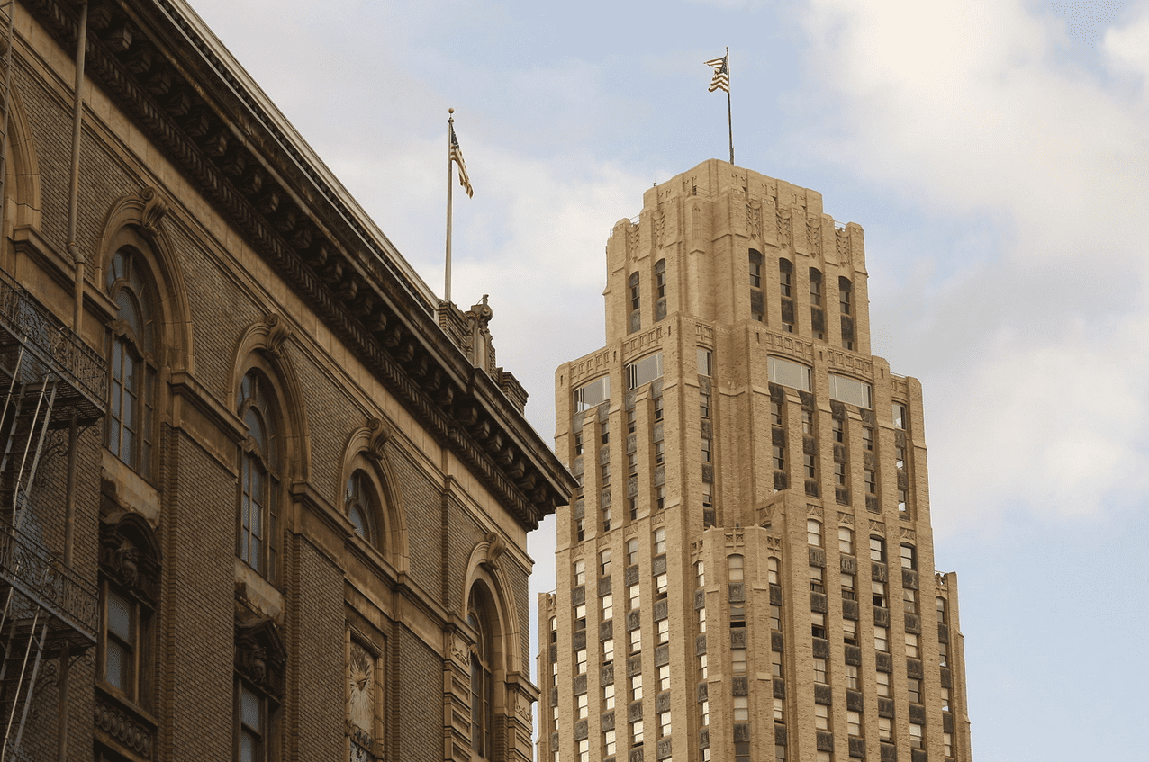 Two buildings next to each other, one darker brown with arched windows. In the background, a tall light yellow building