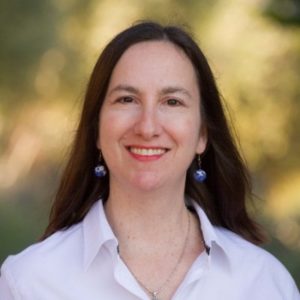 Professor Alice Armitage smiles at the camera in a natural outdoor setting, wearing a white shirt and blue earrings.