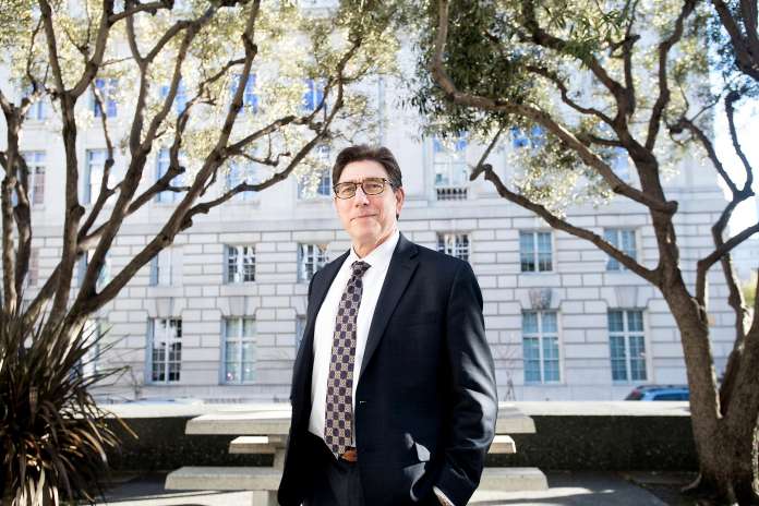 Man wearing glasses and a suit. White building and trees in the background.