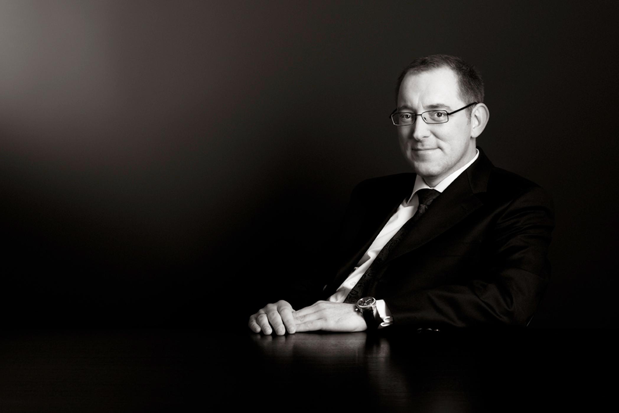 Black and white photo of a man sitting down, wearing glasses and a suit.