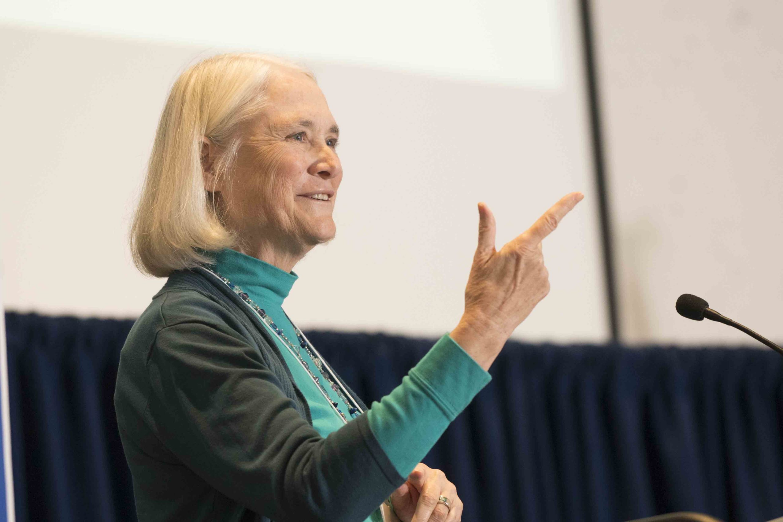 A woman speaks at a podium during a panel discussion, gesturing with her right hand while a microphone is positioned in front of her.