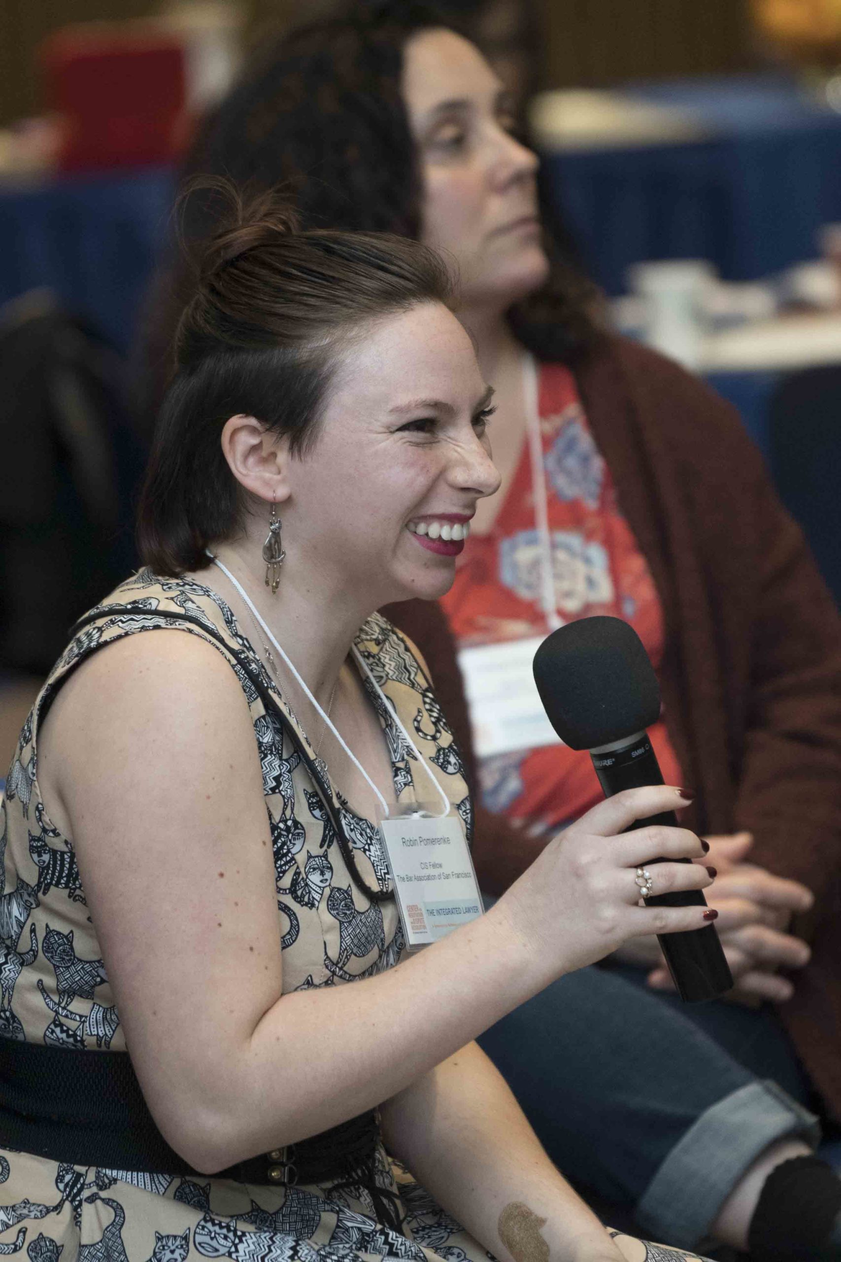 A woman smiles while holding a microphone and speaking during a panel discussion, with another person and table visible in the background.
