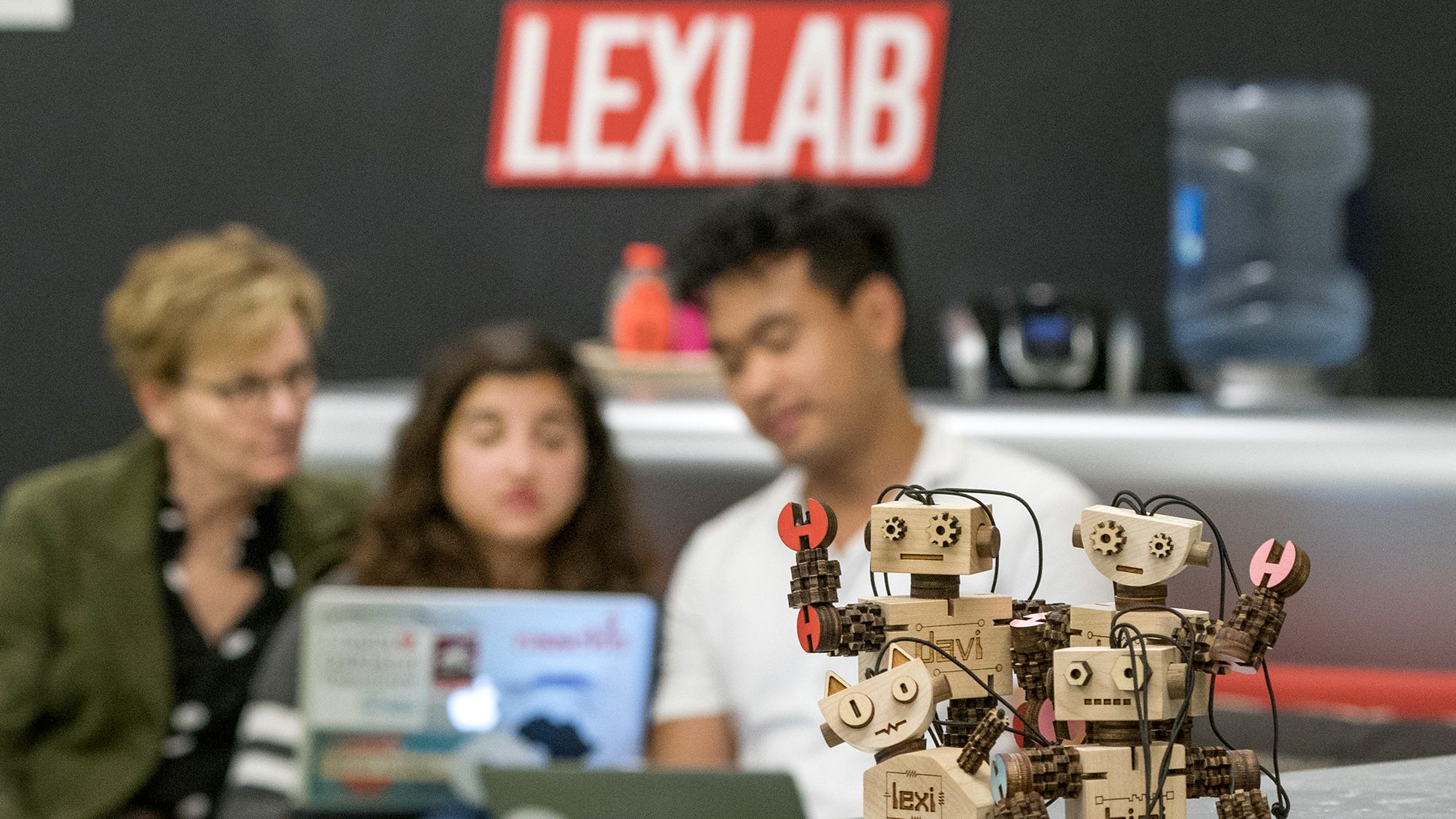 A close-up of three wooden robot figures in the foreground with blurred students working on laptops behind them in a tech-focused environment.