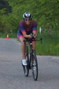 A cyclist in a colorful racing outfit rides a bike on a road lined with greenery. Traffic cones are visible along the route.