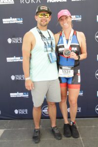 A couple poses for a photo after a triathlon event. The woman holds a finisher's medal and wears a race outfit, while the man stands beside her smiling.