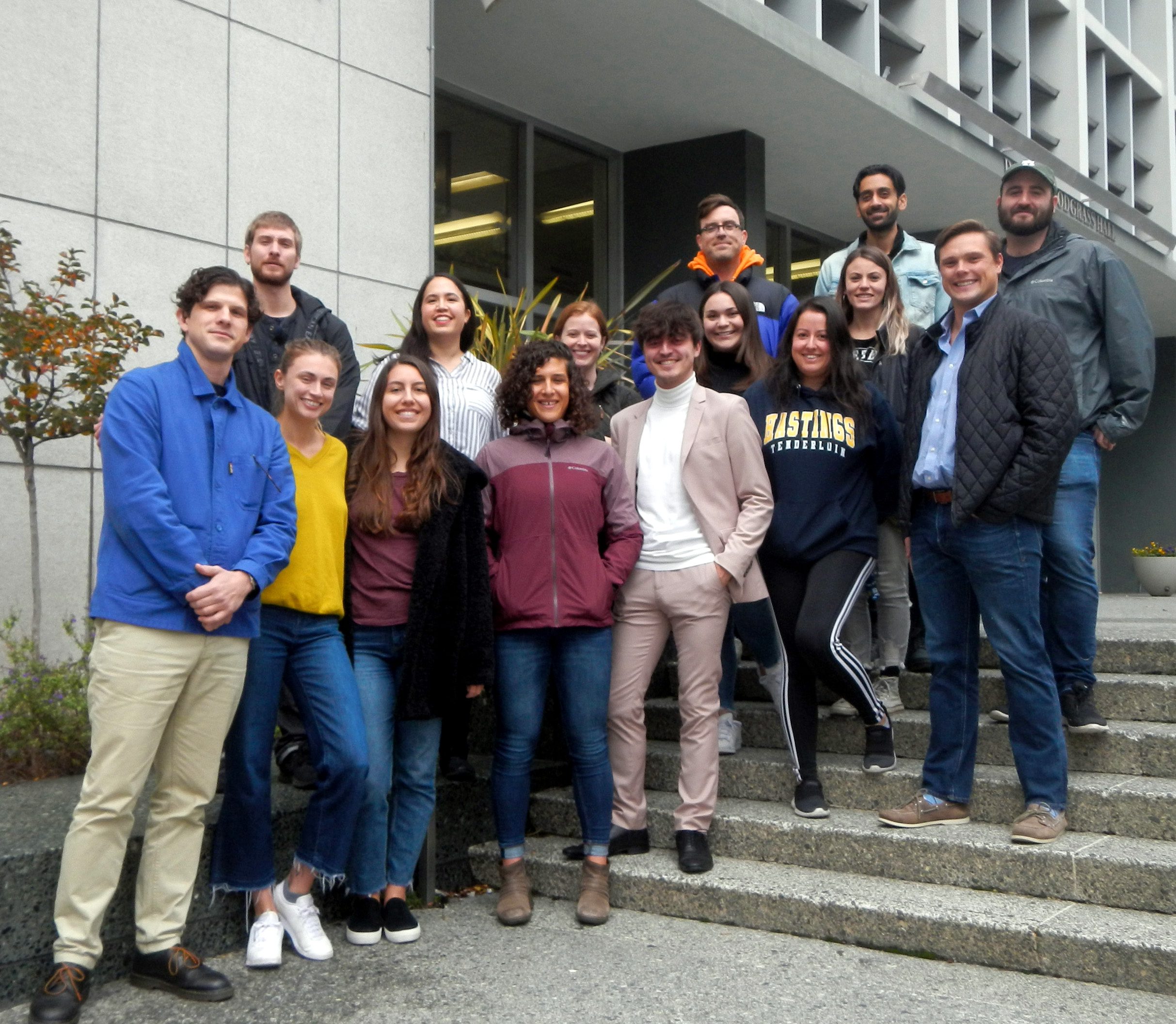 The image shows a group of 17 individuals posing together on steps outside a building, smiling at the camera.