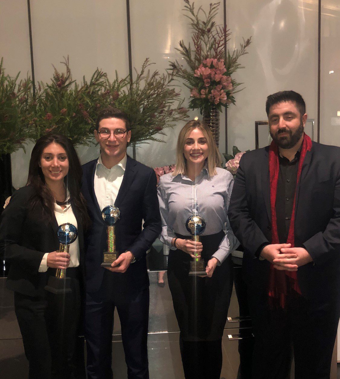 Four individuals are standing together, holding awards, in front of floral arrangements. They are indoors, smiling at the camera.