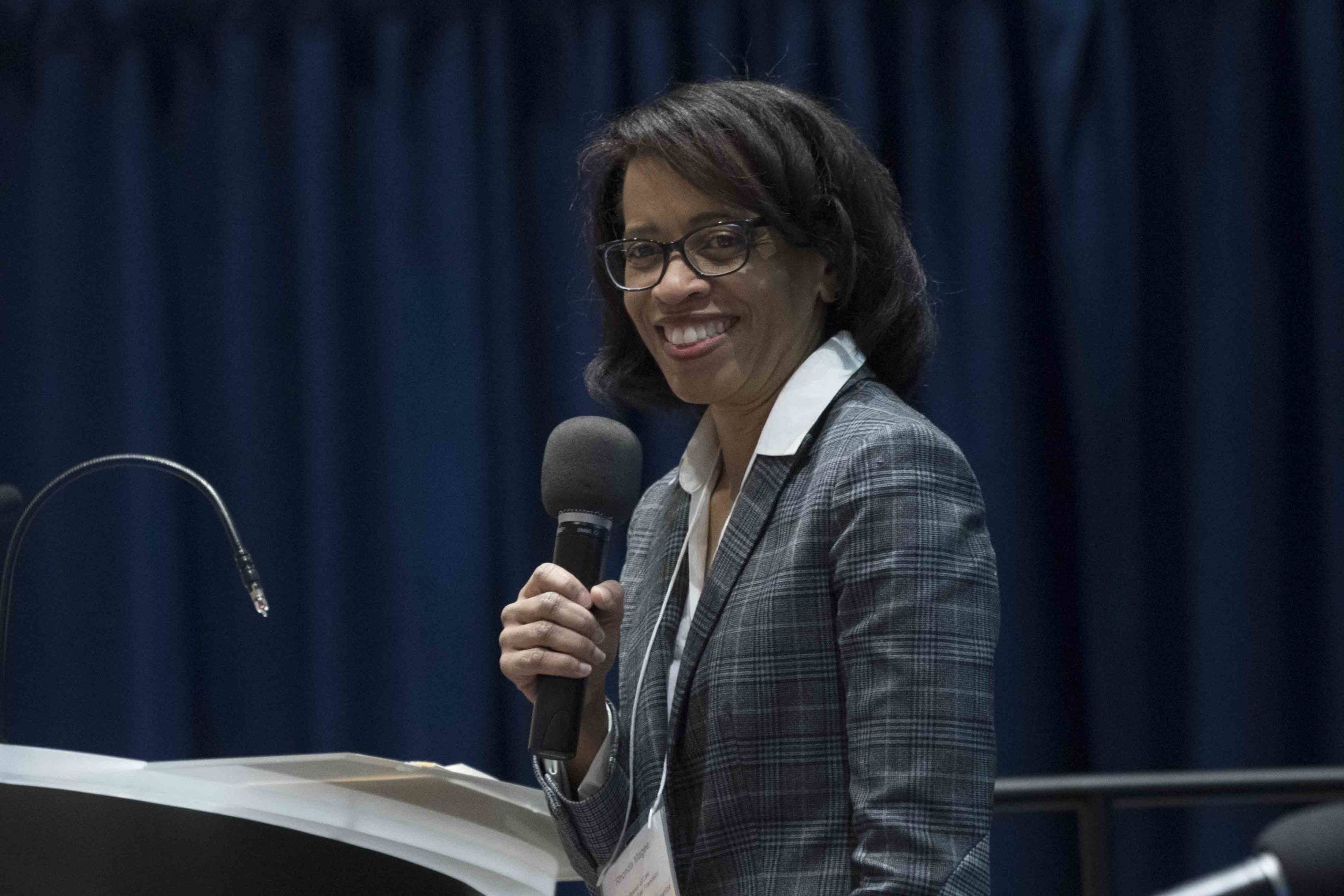 A woman smiles while holding a microphone and speaking at a podium during a panel discussion, with a blue backdrop in the background.