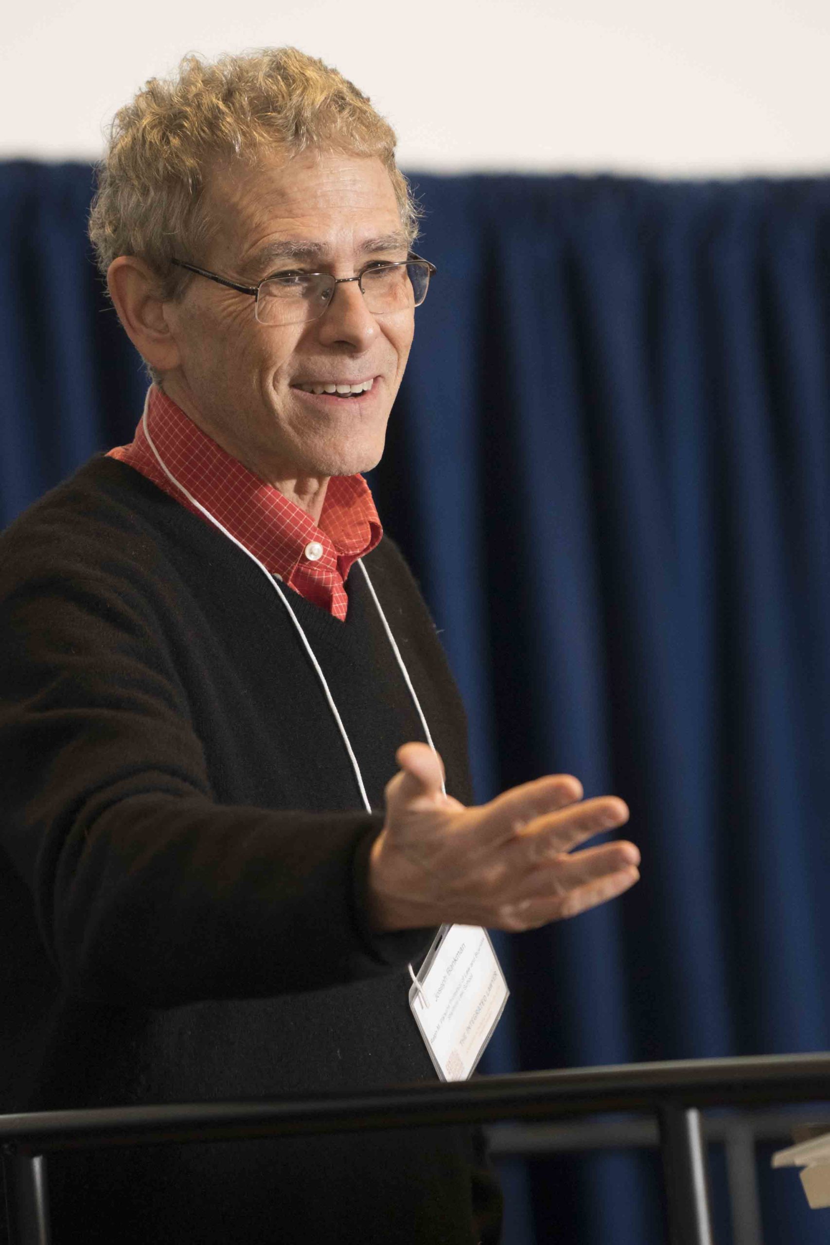 A man wearing glasses and a black sweater gestures while speaking, with a blue backdrop and a table visible in the foreground.
