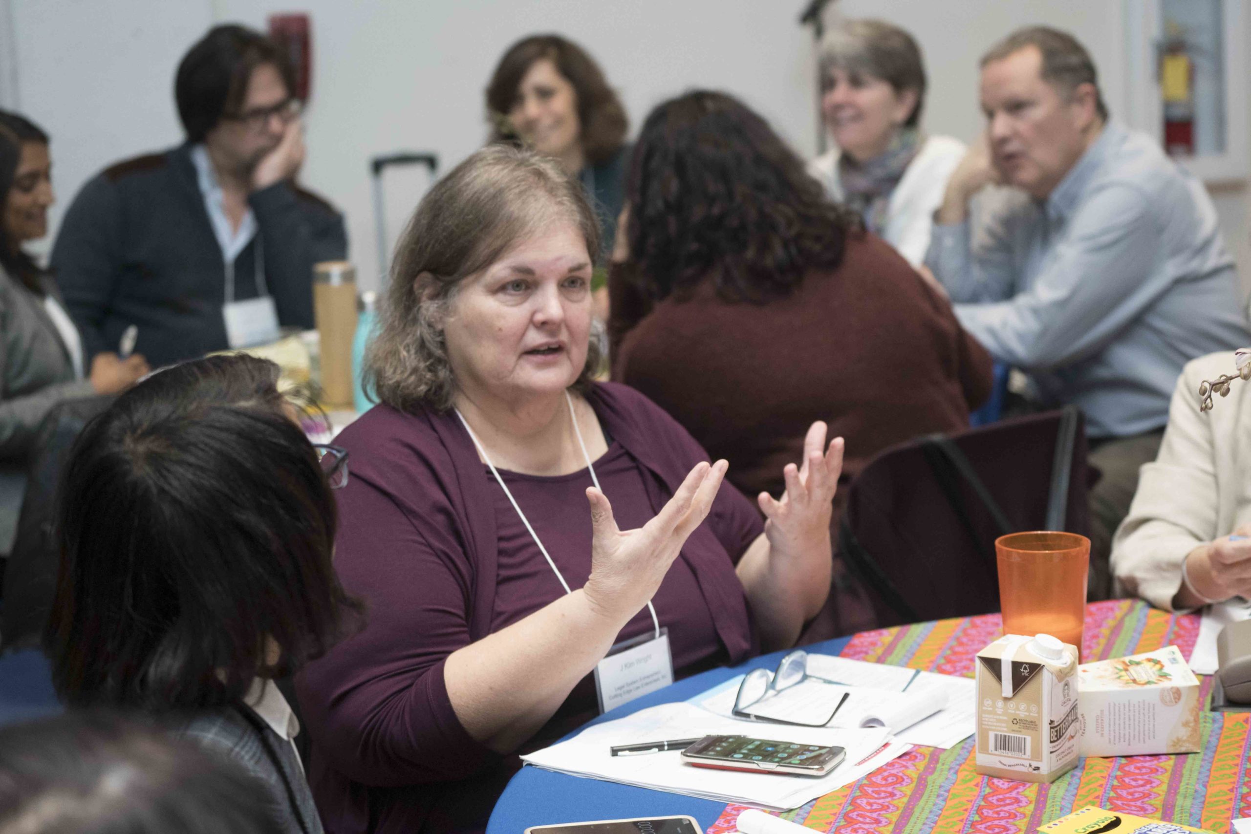 A woman gestures while speaking at a table during a discussion, with several people seated in the background. Papers and a phone are visible on the table.