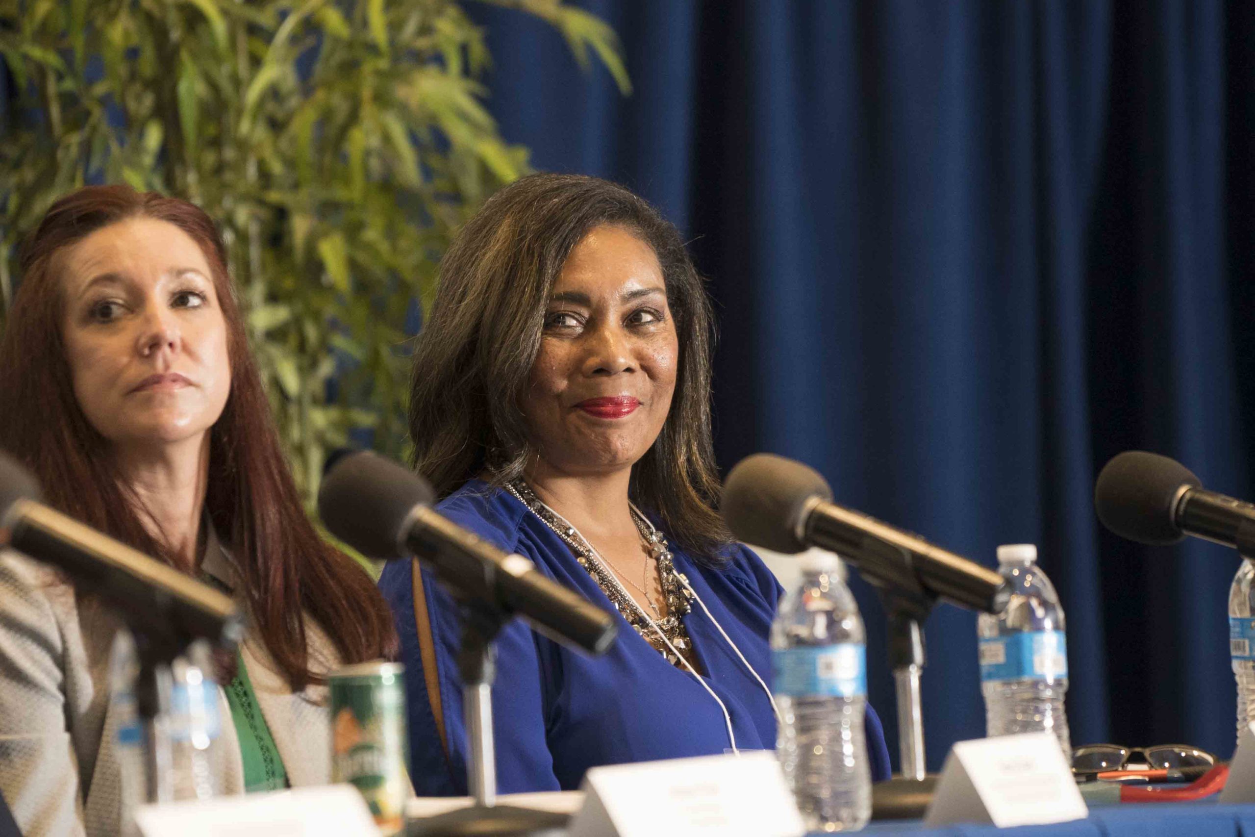 A panel discussion featuring two women seated at a table with microphones and water bottles, with greenery in the background.