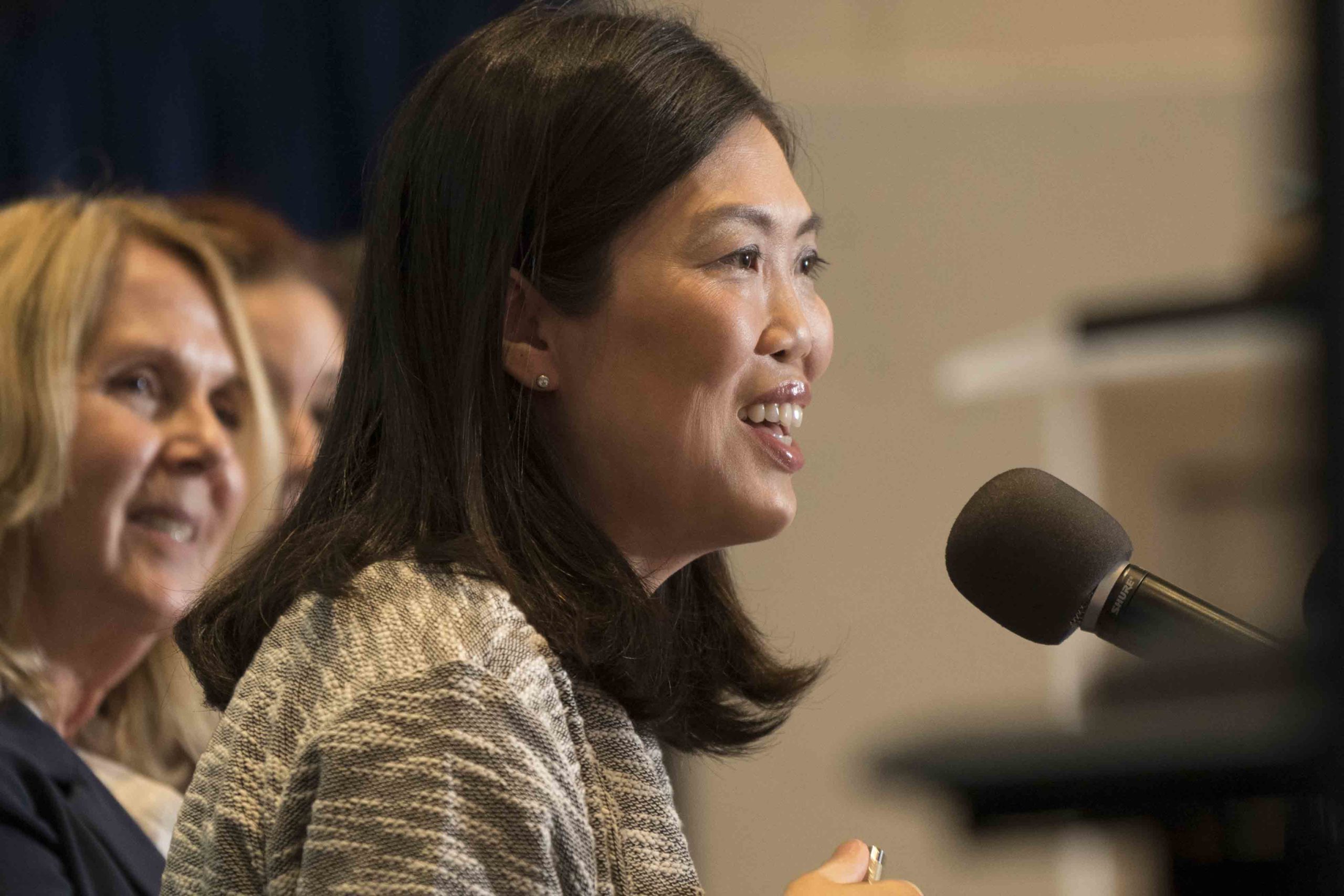 A panel discussion featuring a woman speaking into a microphone, with two women in the background, all seated at a table.
