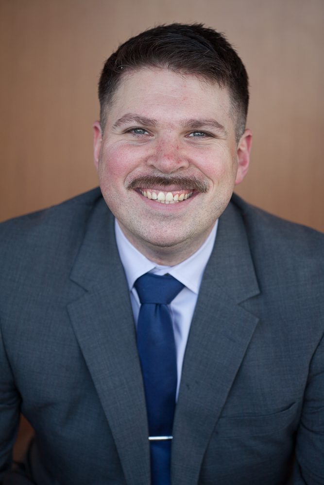 Aaron Chase, a law student, smiles while wearing a suit and tie, posing for a professional portrait against a wooden background.
