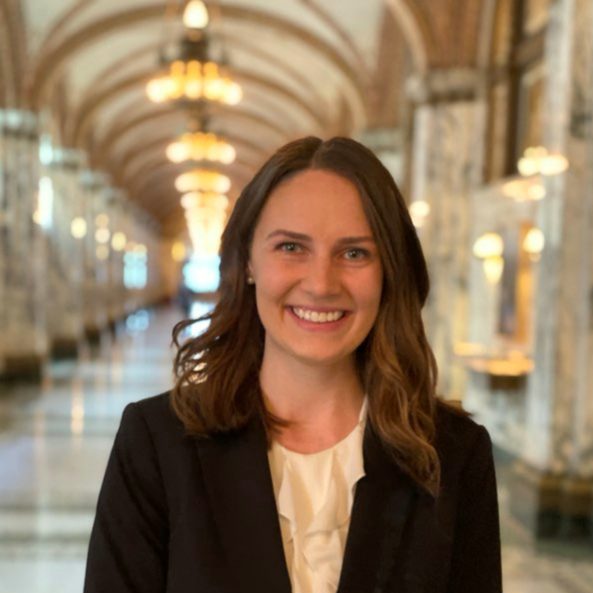A smiling individual in professional attire stands in an elegant hall with arches and chandeliers in the background, likely within a government building.
