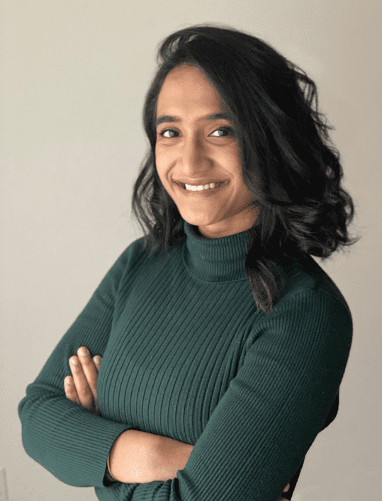 A student smiles with arms crossed, wearing a green turtleneck sweater, against a light-colored background in a professional setting.