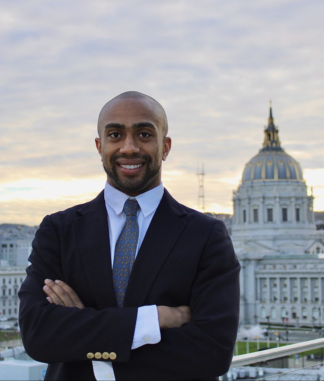 A man in a suit smiles with his arms crossed, standing against a backdrop of a cityscape featuring a government building under a cloudy sky.