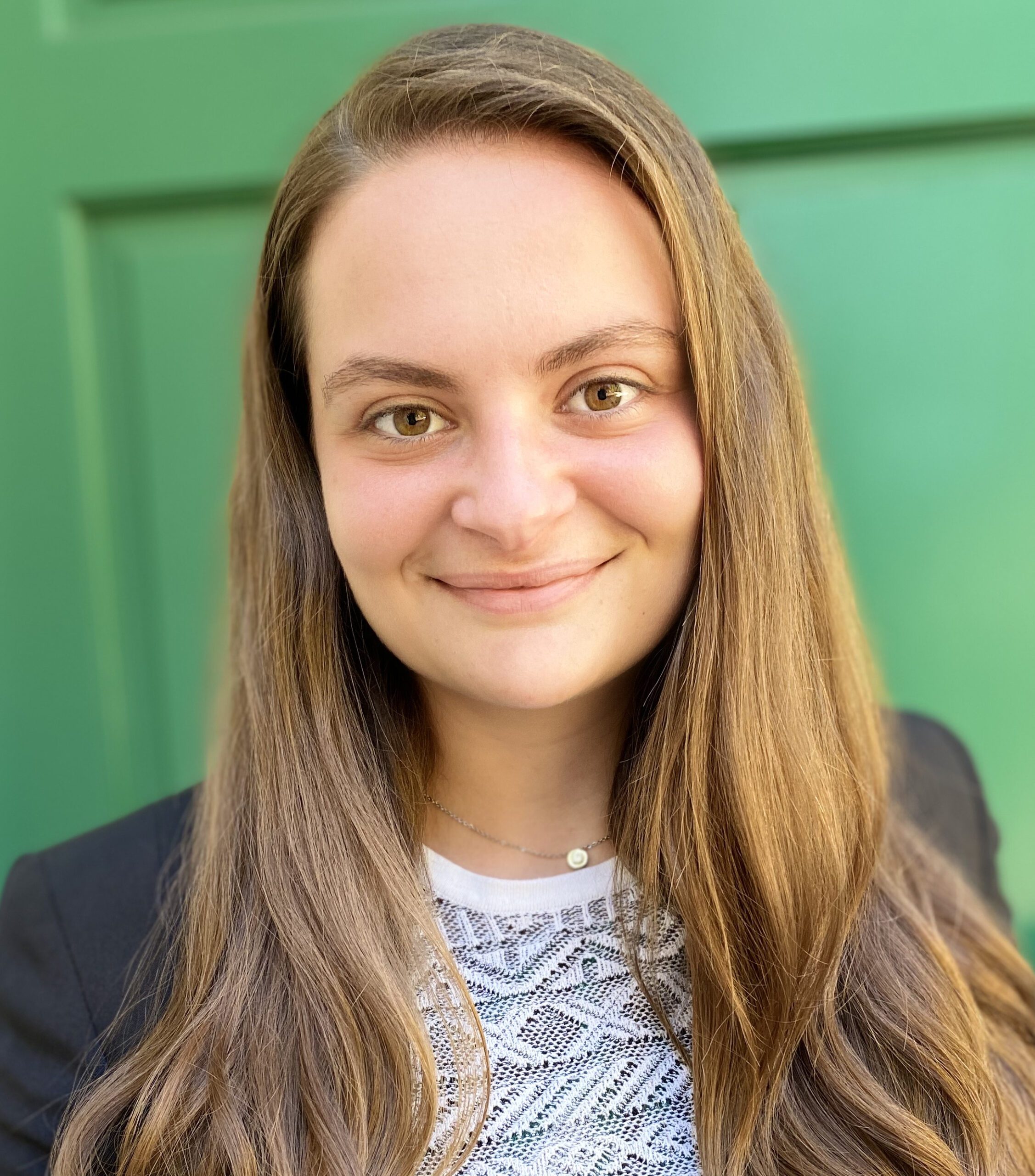 Maddy Klein smiles in front of a green background. She is wearing a black blazer over a white top, her long hair falls naturally.