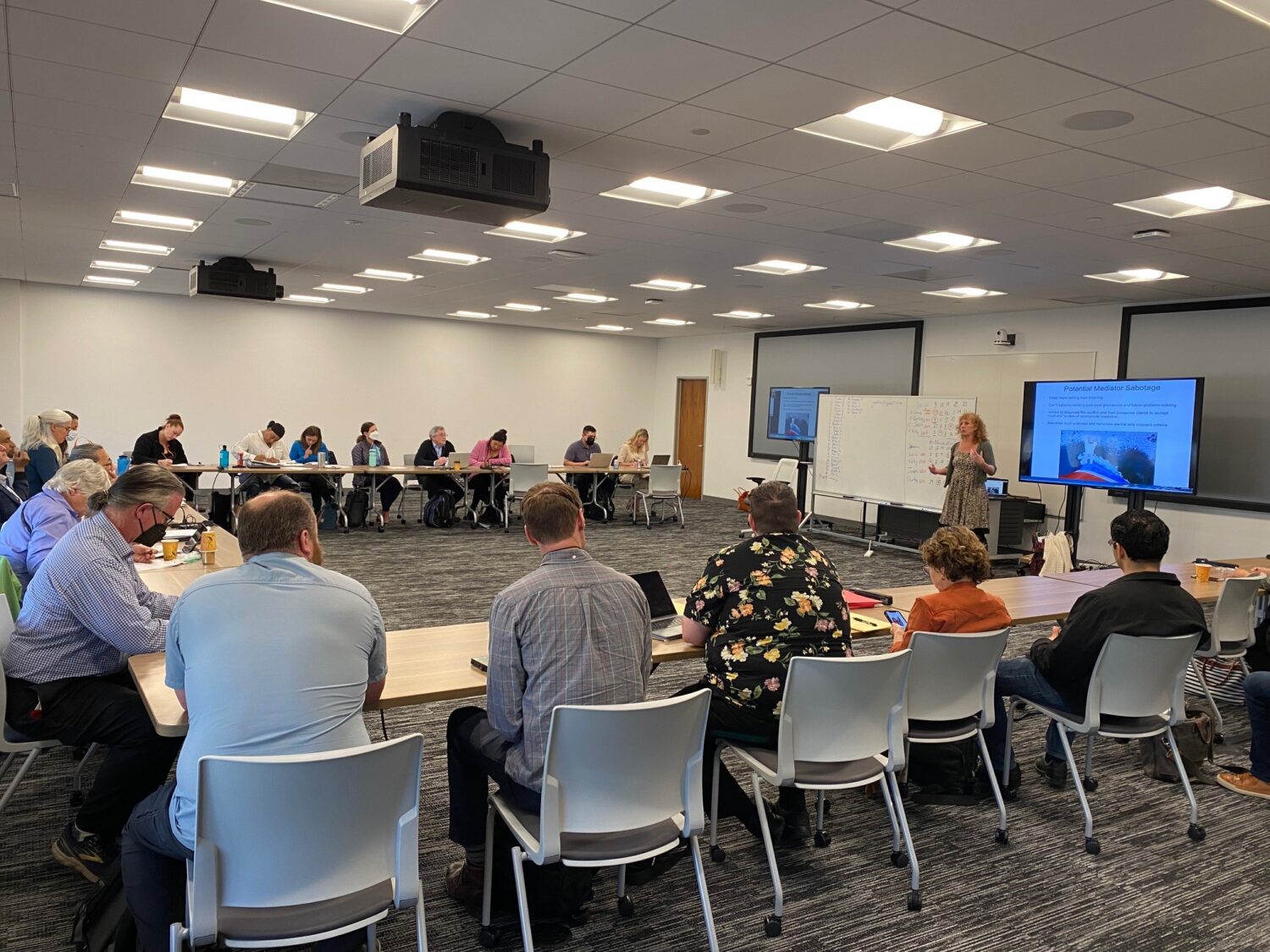 A group of participants engaged in a mediation training session. A presenter stands at the front, while attendees observe and take notes.