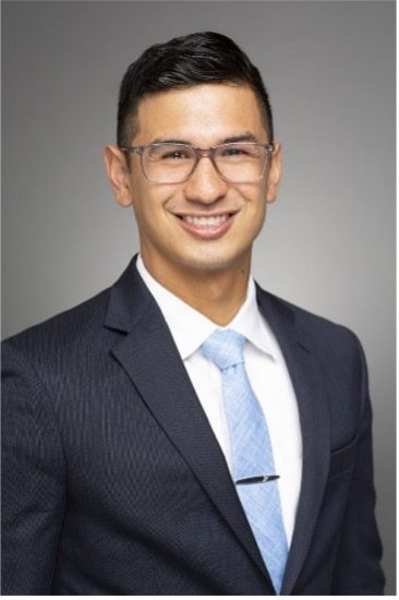 Andrew Angeles, an attorney for BART, smiles in a professional suit and tie, with a light blue tie and glasses, against a gray background.
