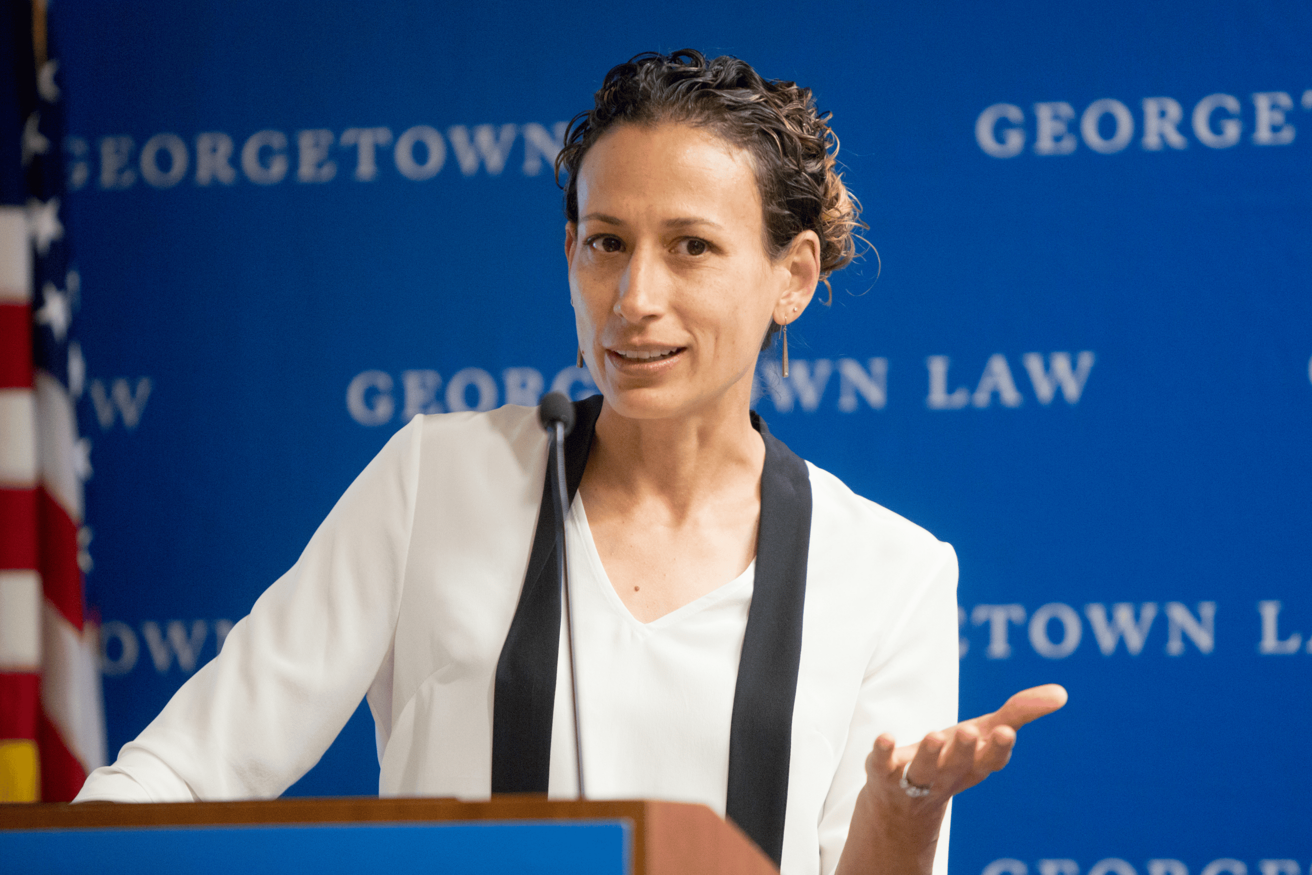 Professor Thalia González speaks at a podium during an event, gesturing with her hand and engaged with the audience around her.