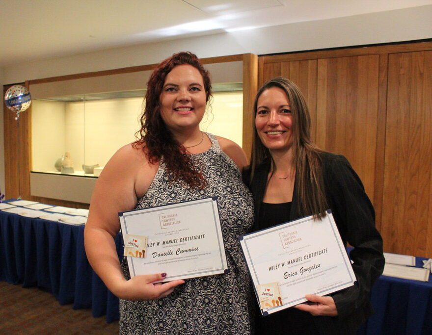 Danielle Cummins and Erica Gonzales hold their Wiley W. Manuel Certificates at a public interest awards ceremony, smiling in front of a display table.