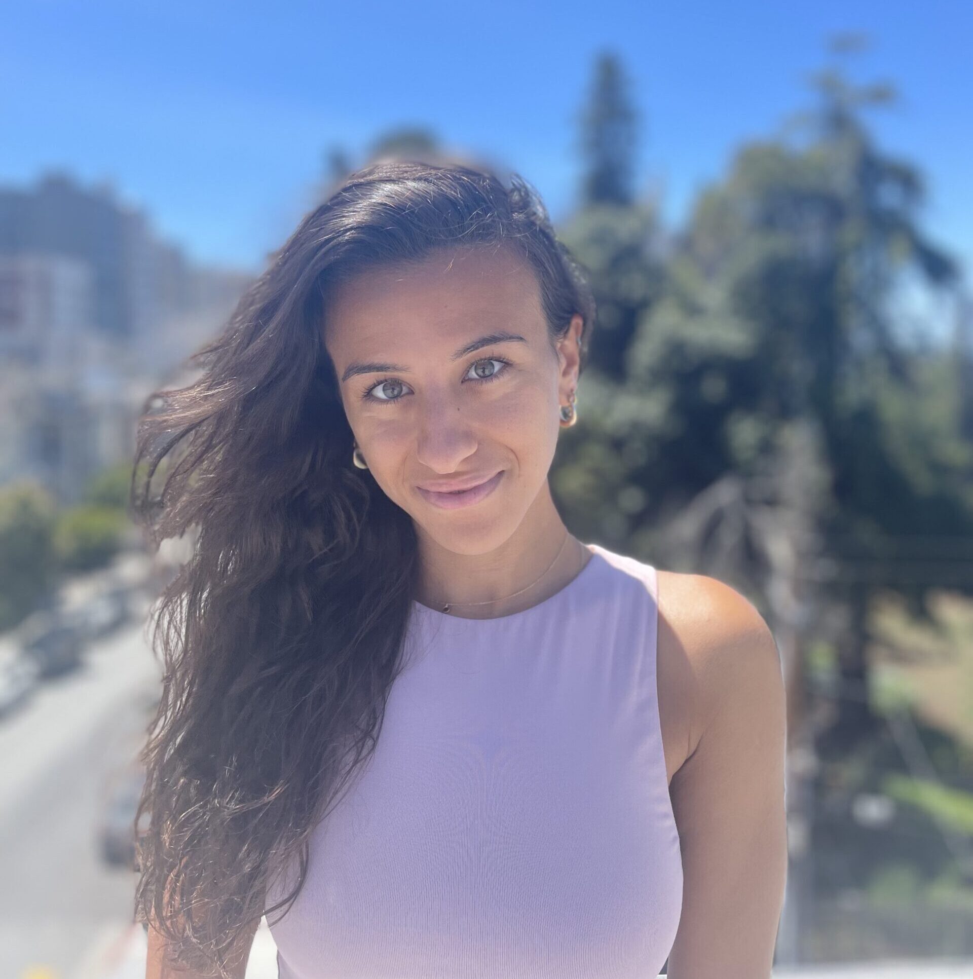 Yasmine Hajjaji poses outdoors with a clear blue sky and trees in the background, smiling while wearing a light purple sleeveless top.