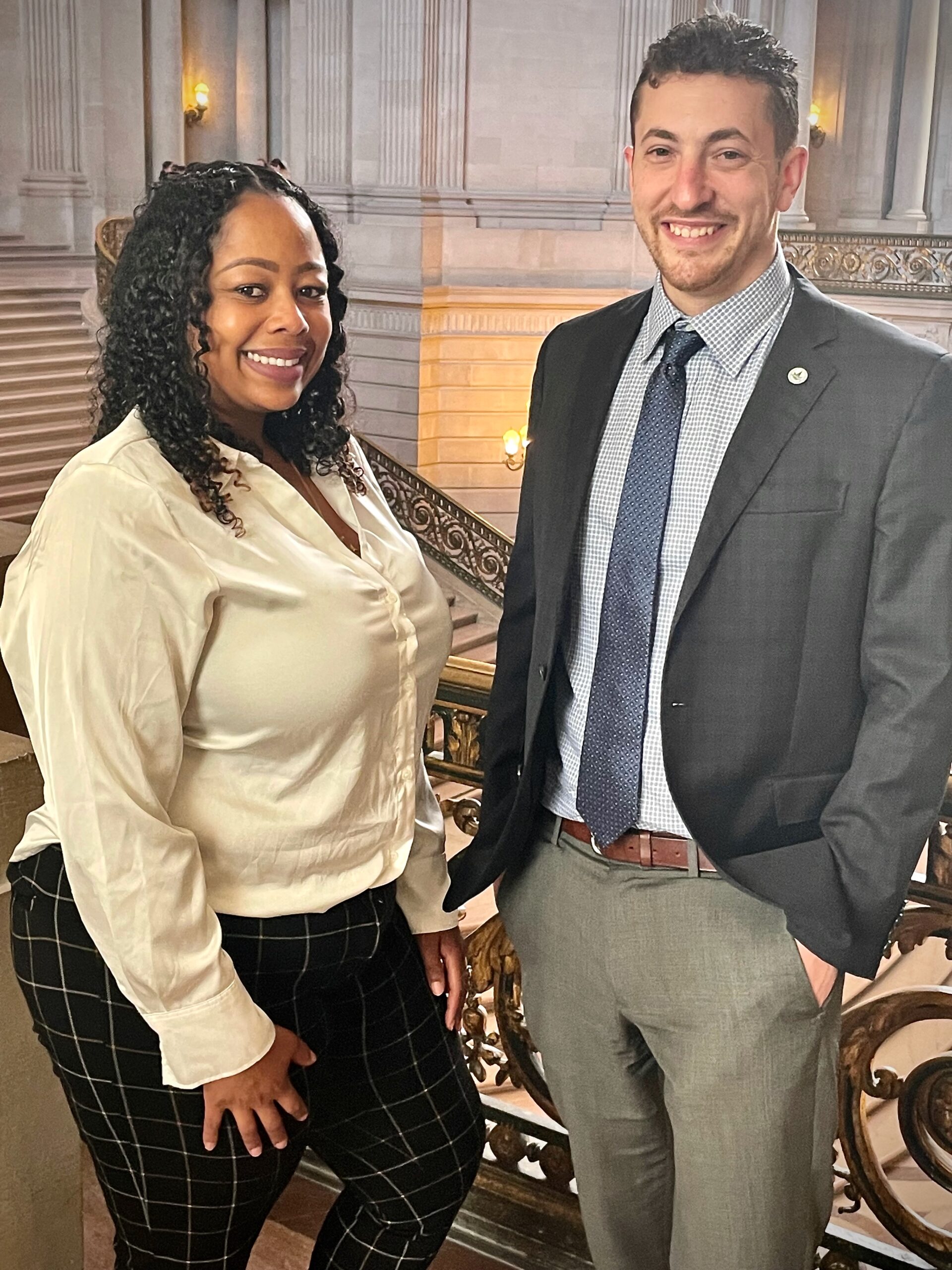 Two professionals stand together in a historic building, smiling. One wears a white blouse and patterned pants, while the other is dressed in a suit.