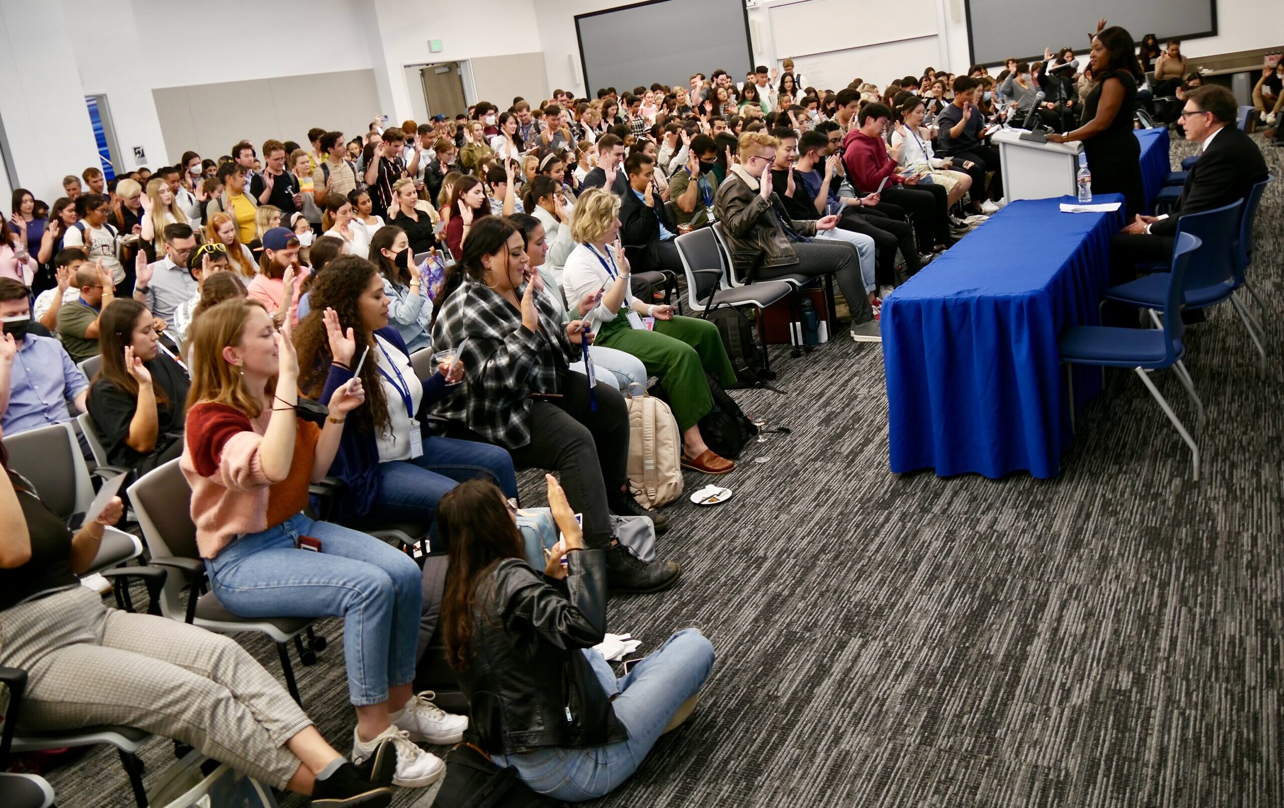 A large group of students in a lecture hall raise their hands during a professionalism pledge at a convocation ceremony.