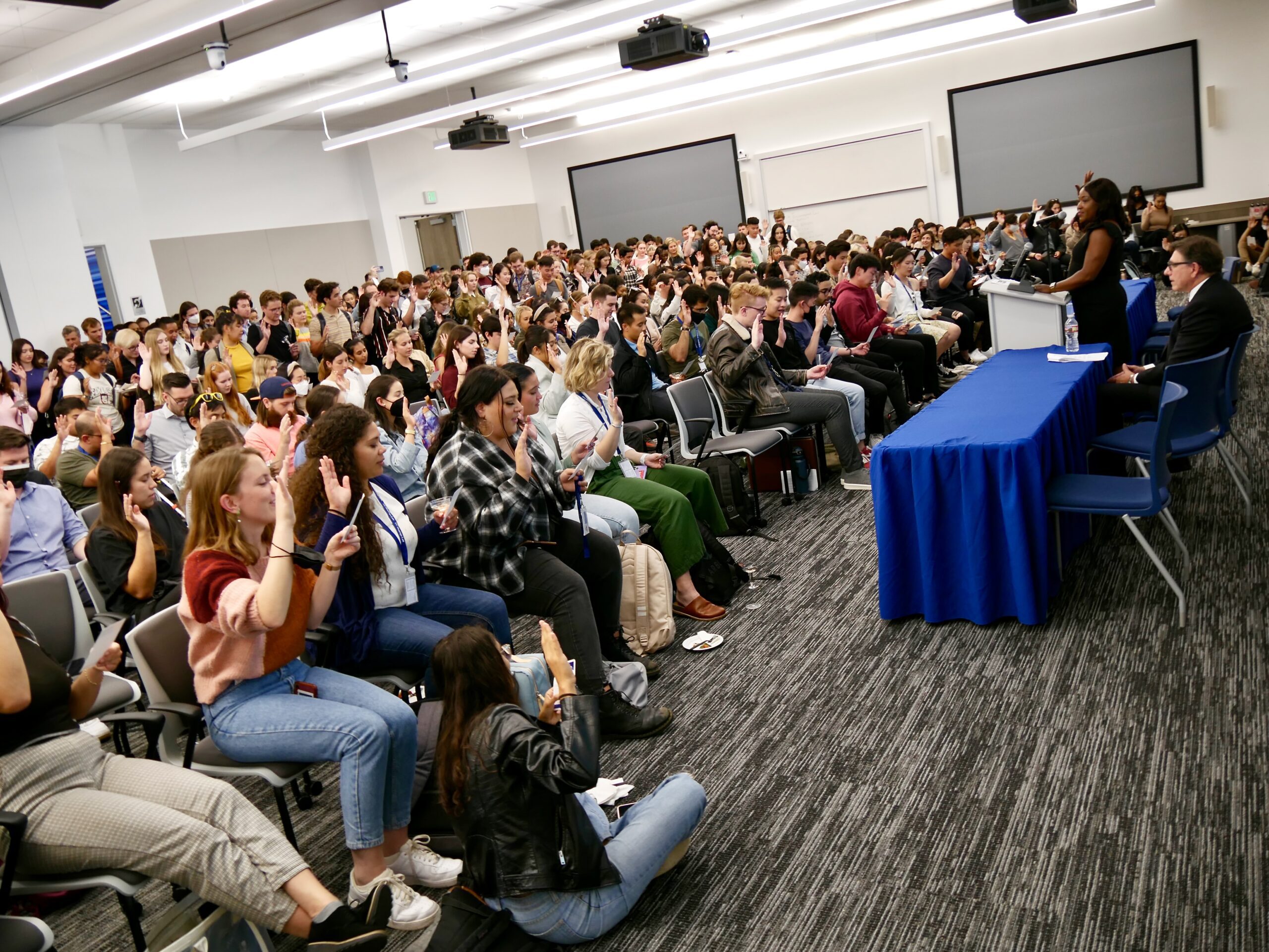 A large group of students in a lecture hall raise their hands during a professionalism pledge at a convocation ceremony.