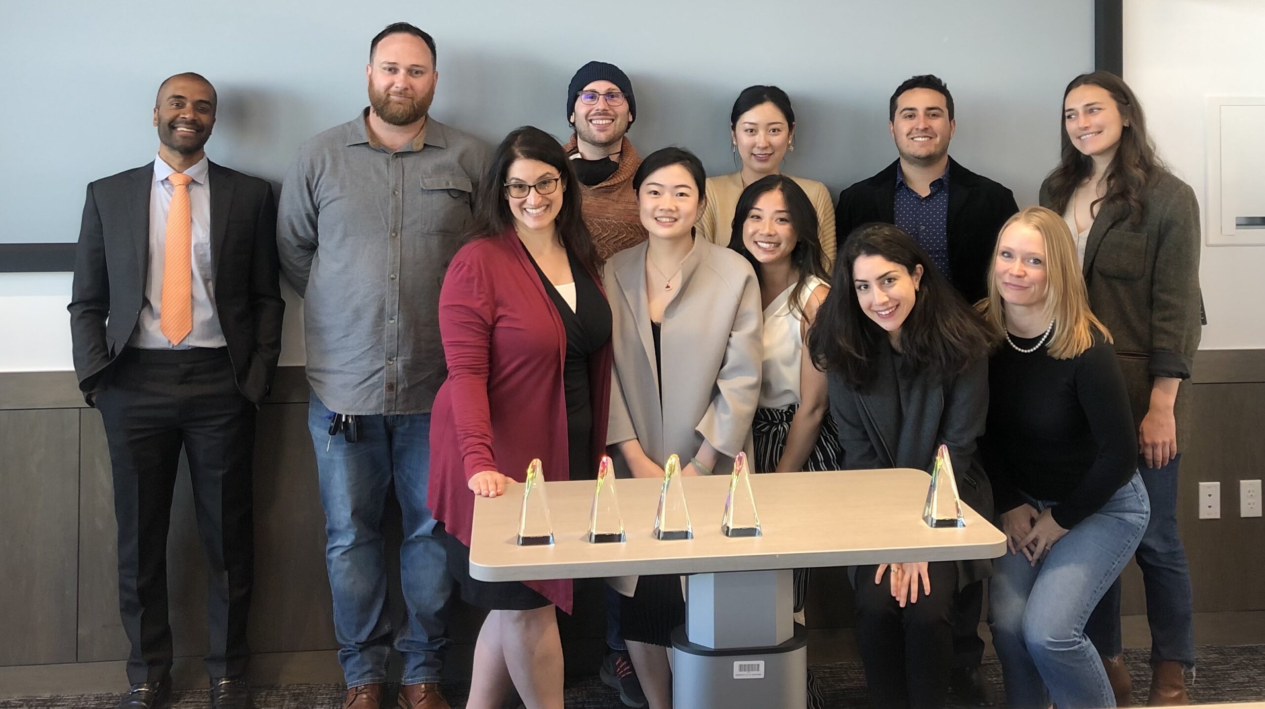 A group of twelve individuals poses together at a graduation and awards ceremony, featuring four trophies on a table in front of them.