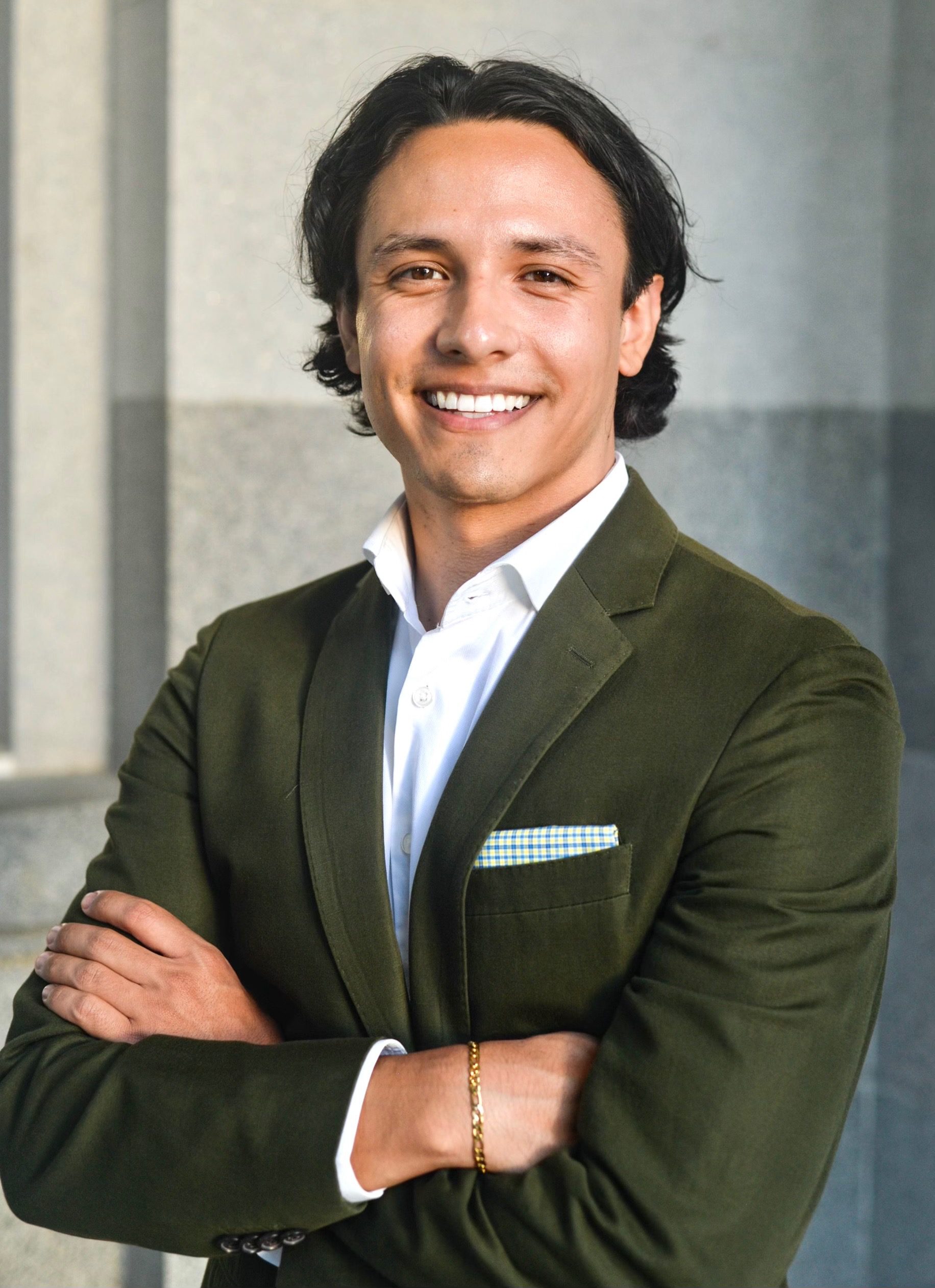UC Law SF student Victor Torres smiles while wearing a dark green blazer over a white shirt, with arms crossed against a gray background.