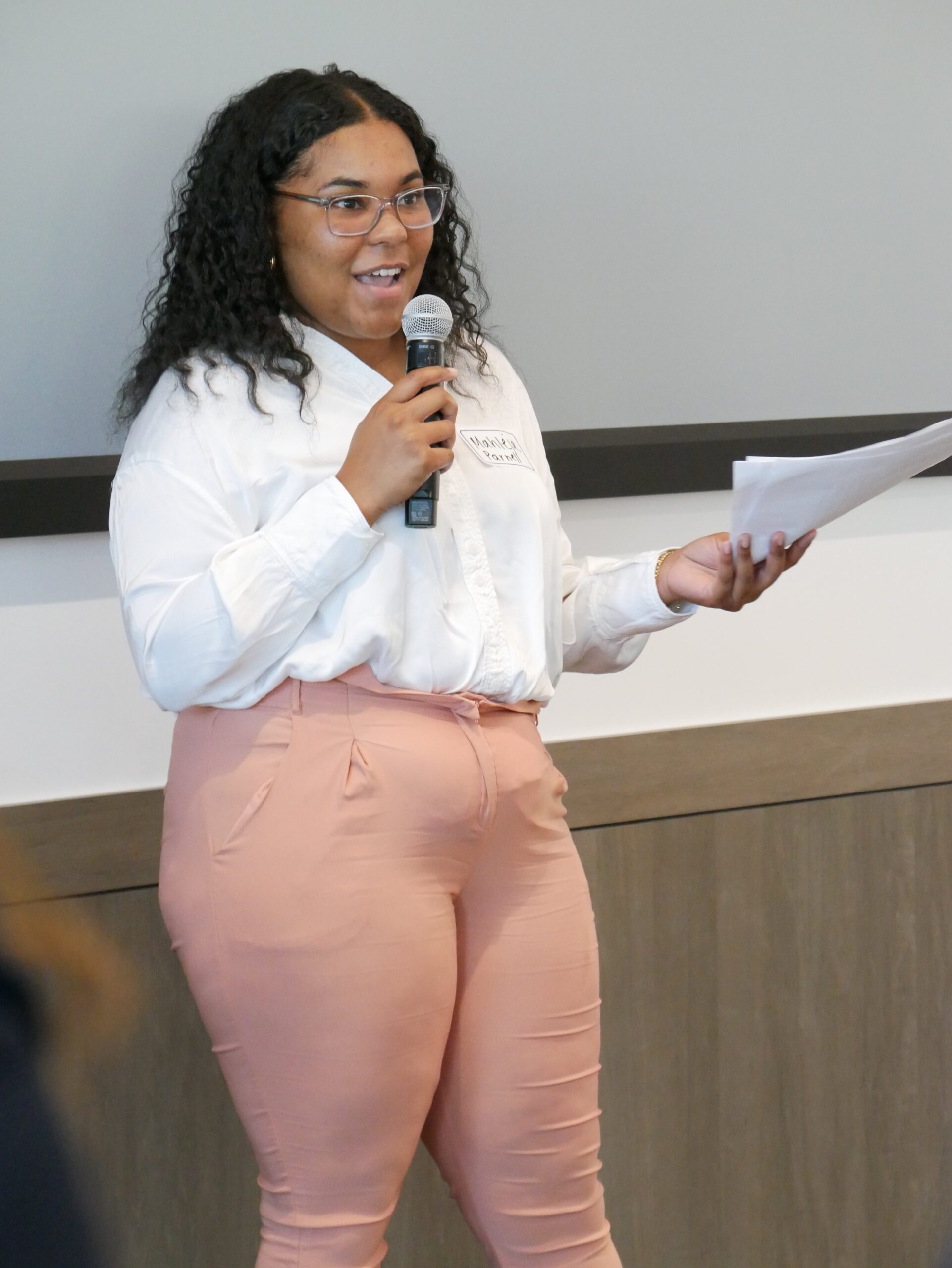 Mahleja Parnell stands at a podium, speaking into a microphone, holding papers in her hand. She is wearing a white shirt and pink trousers.