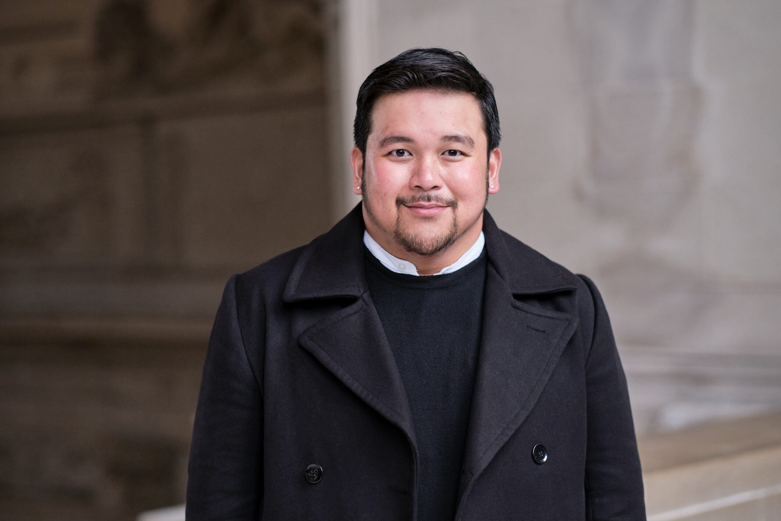 UC Law SF student Alex Allam smiles while wearing a black coat and a sweater, posing outdoors in a historic setting.
