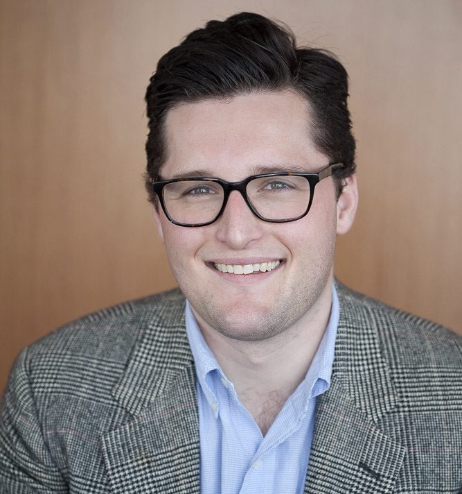 Alexander Vicas smiles for the camera, wearing a plaid blazer over a light blue collared shirt, against a wooden backdrop.