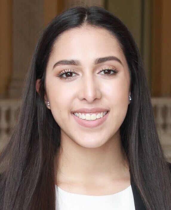 Adriana Diaz Mireles smiles for a headshot in a formal setting, wearing a black blazer and a white top, with long dark hair and earrings.