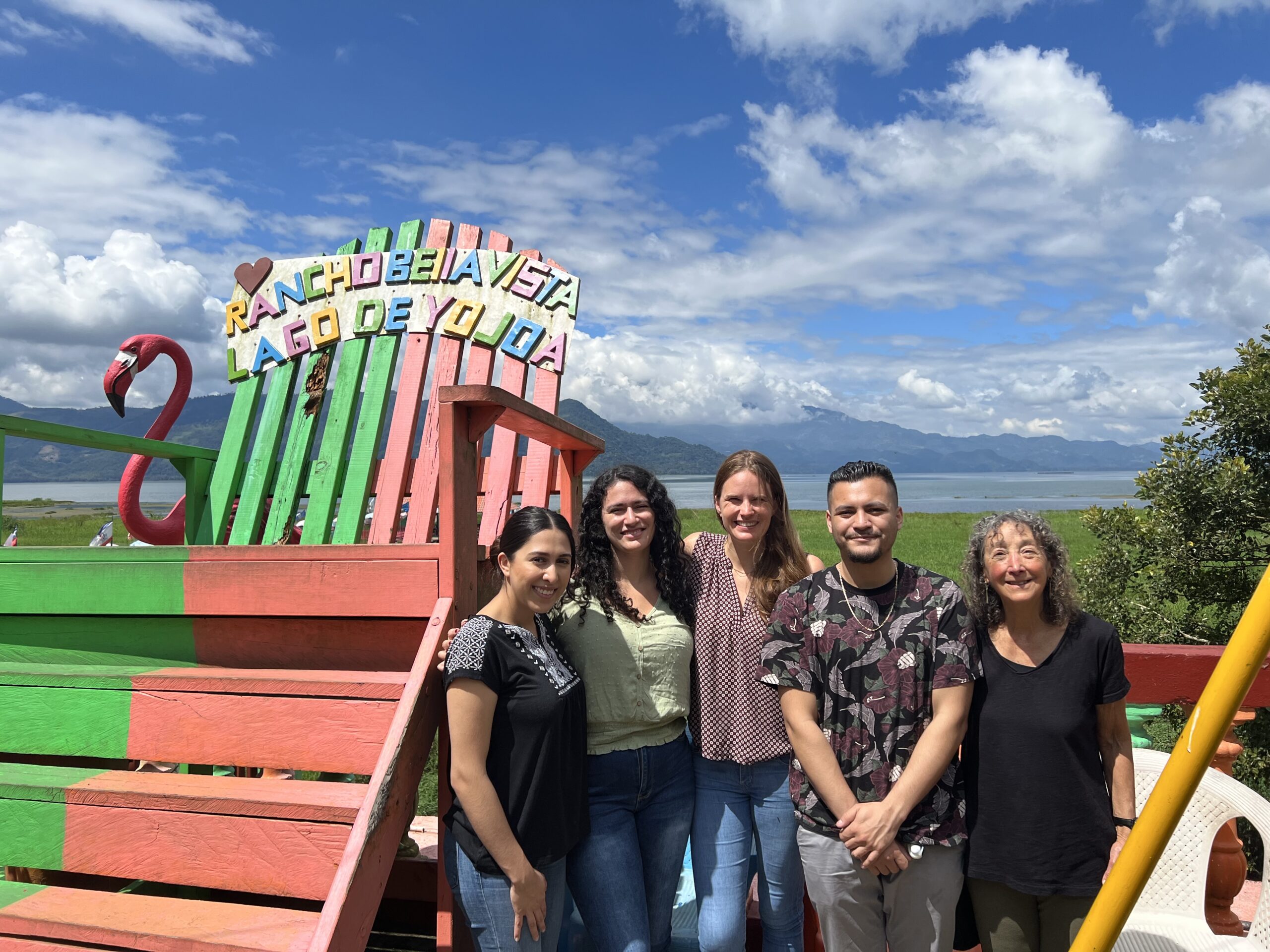 Five individuals pose together in front of a colorful seating area with a sign that reads "RANCHO BEIJAVISTA LAGO DE YOJOA." A scenic lake and mountainous background is visible.