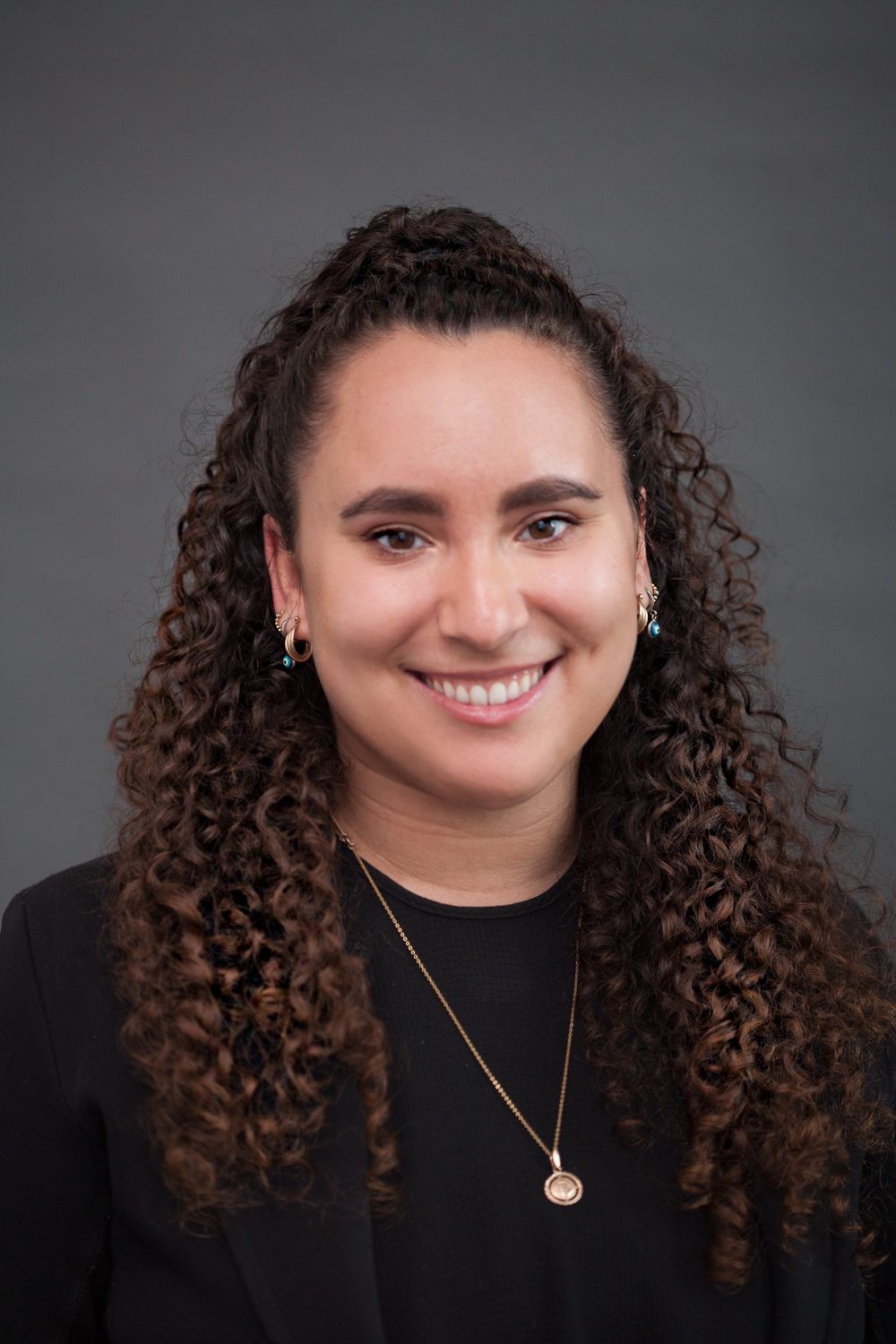 Melanie Gorelik smiles at the camera, wearing a black top and a necklace, with curly hair cascading down her shoulders against a neutral background.