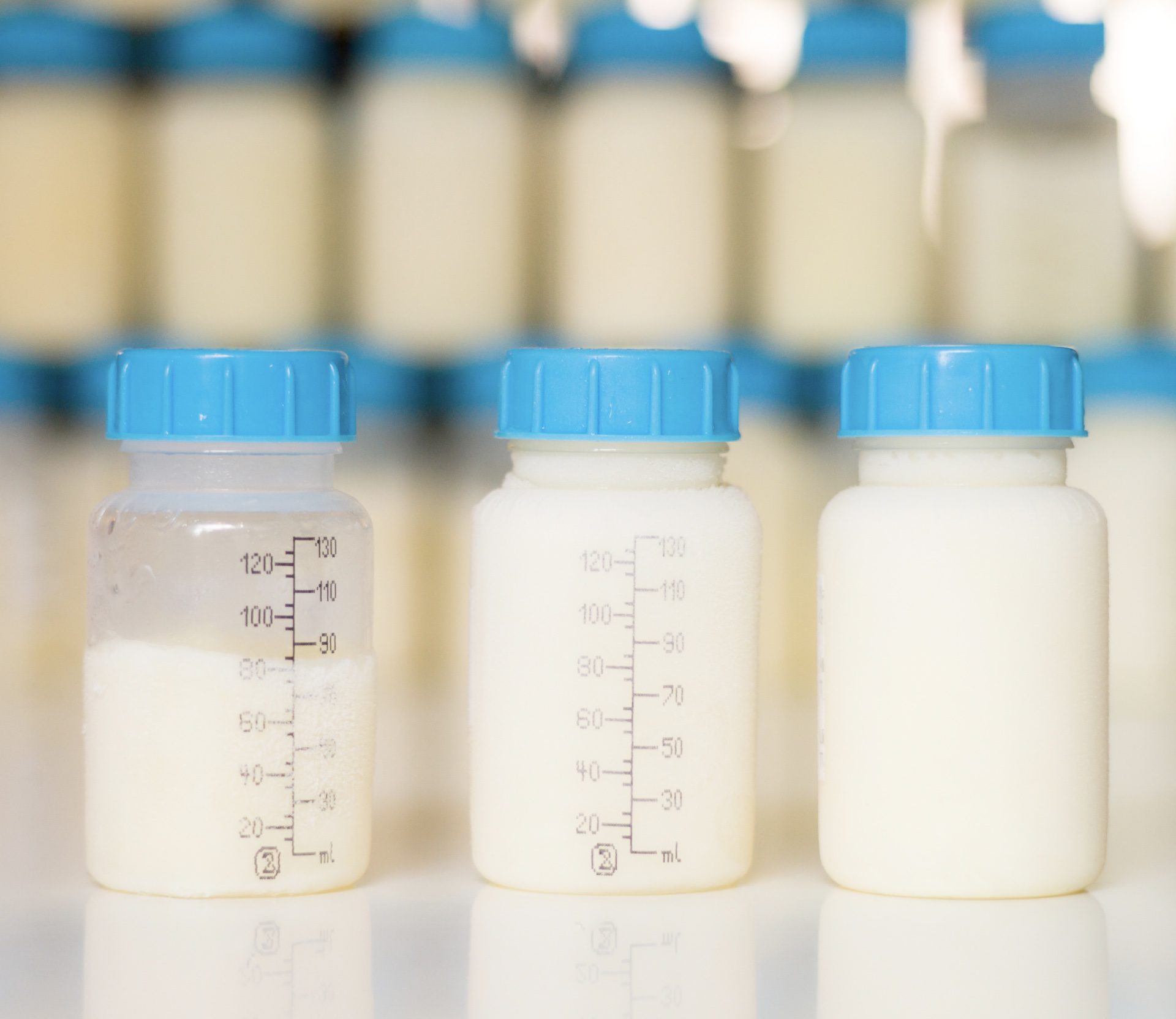 Three baby bottles with blue caps, filled with a dark liquid, a cloudy liquid, and a white liquid, are arranged in a row on a reflective surface.