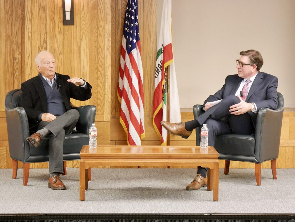 George Roberts and David Faigman sit in armchairs, discussing legal education and investment during a fireside chat at UC Law SF, flanked by flags.