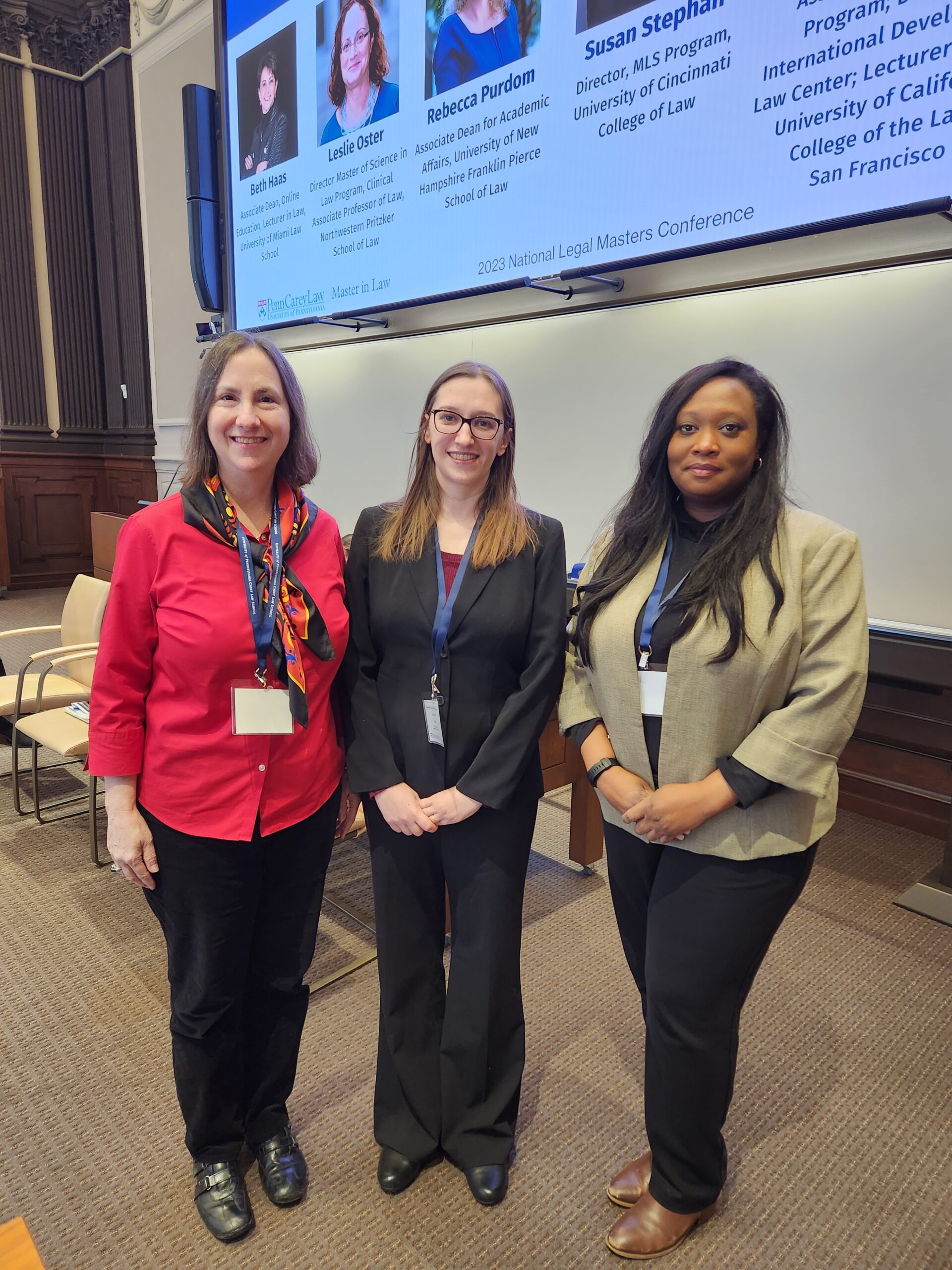 Three women pose together at a conference, displaying name tags. A presentation is projected on the screen behind them.