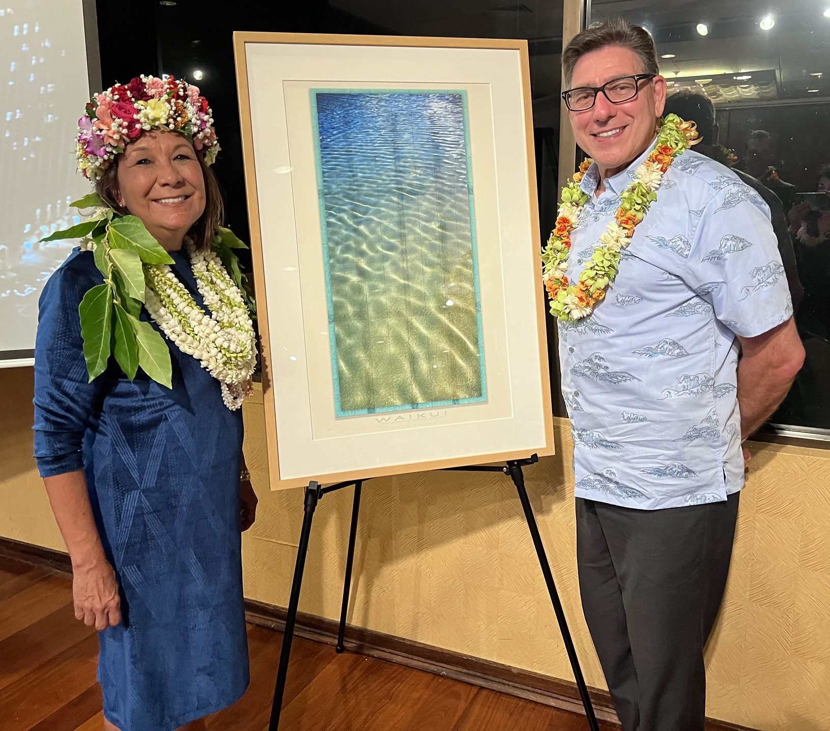 Crystal Rose and David Faigman stand together, smiling with leis, beside an artwork depicting water. The setting is a gala event in Hawaii.