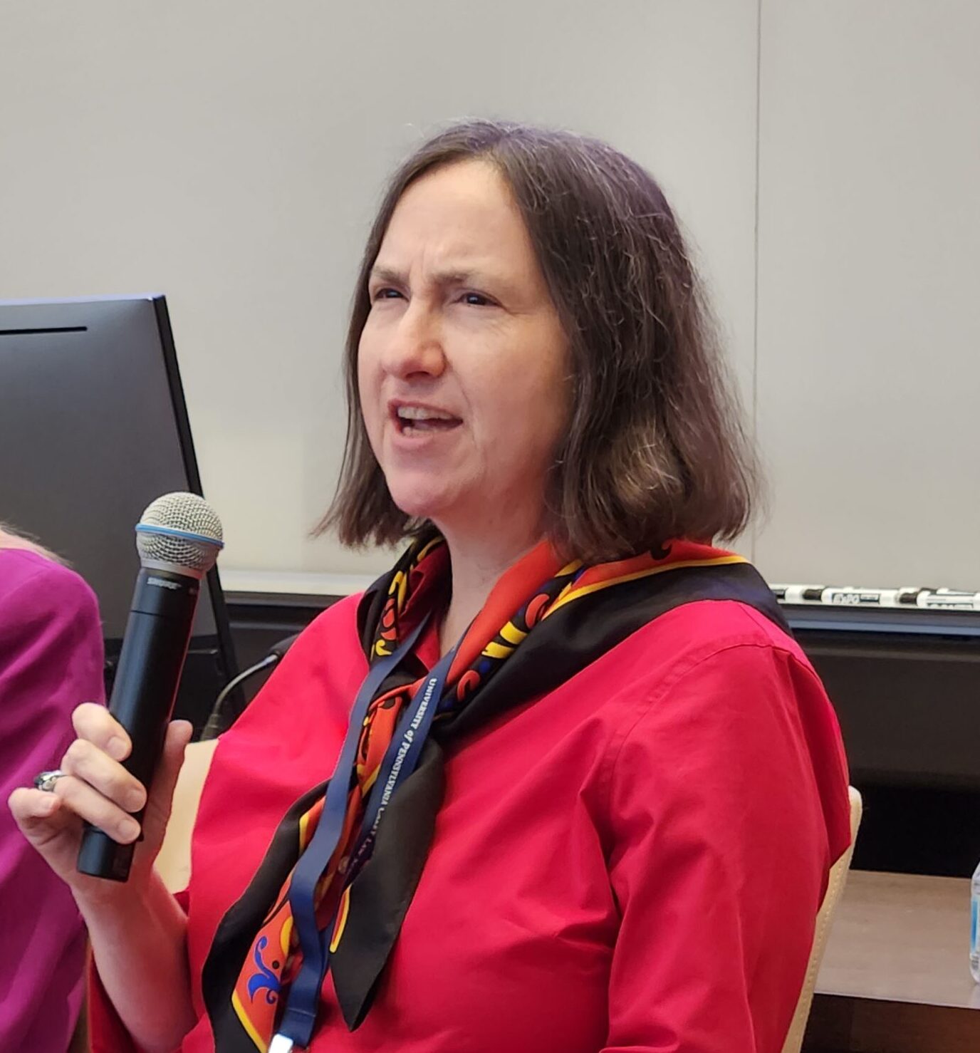 Jessica Vapnek speaks into a microphone during a panel discussion at the Legal Masters Conference. She is wearing a red shirt and a patterned scarf.