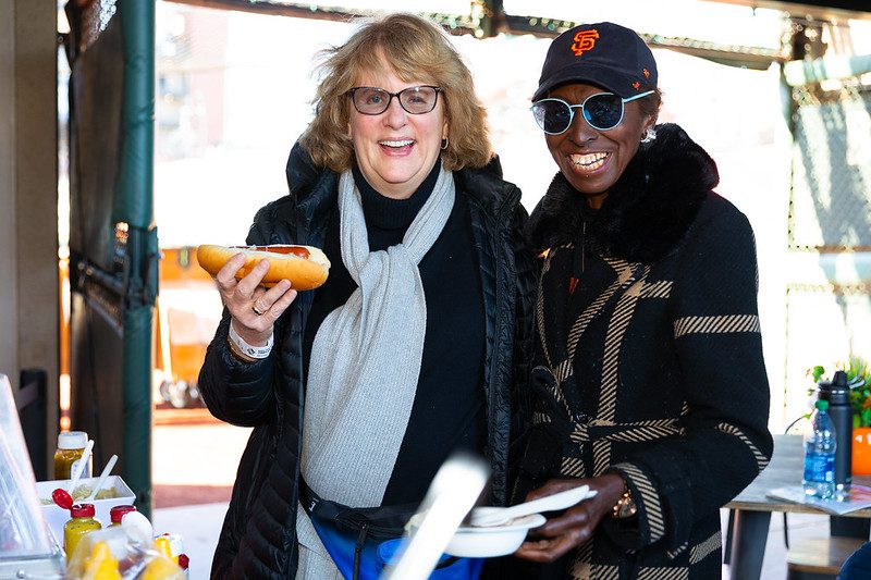 Two women smile for the camera while holding food at Oracle Park, with condiments and drinks visible on the table in front of them.