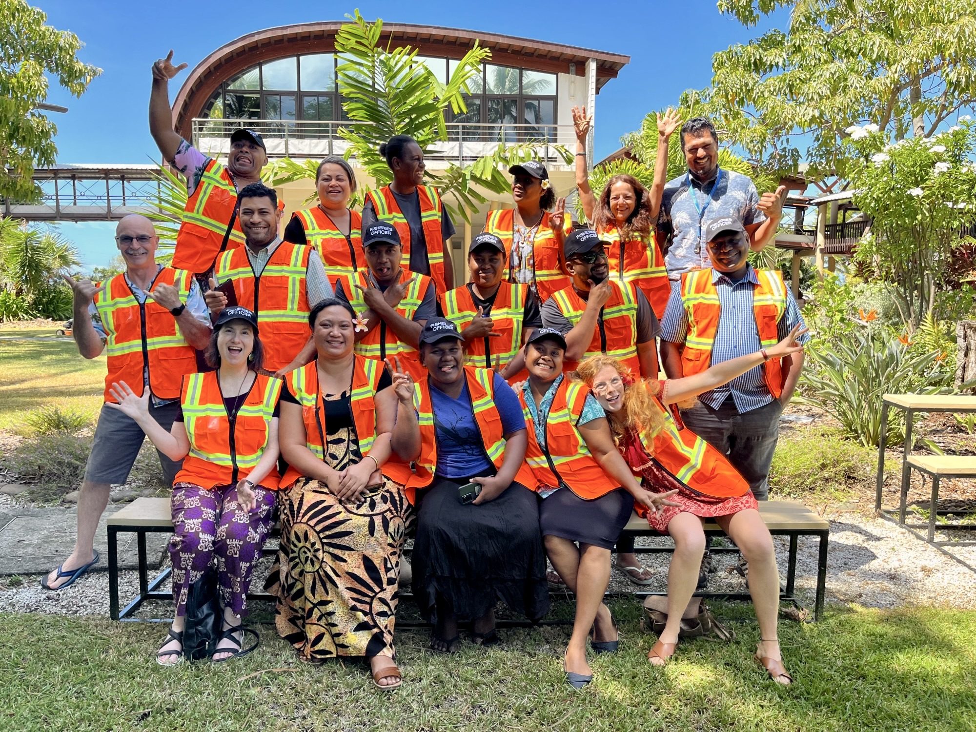 A diverse group of individuals in bright safety vests poses for a photo outdoors, smiling and making gestures, with a tropical background.