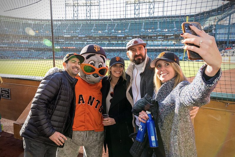 A group of four adults poses for a selfie with a San Francisco Giants mascot at Oracle Park during a baseball event.