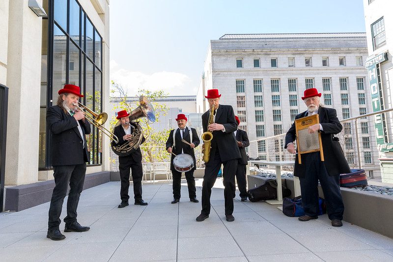 A jazz band of six musicians wearing black suits and red hats performs outdoors, with instruments including a trombone, saxophone, and drum.