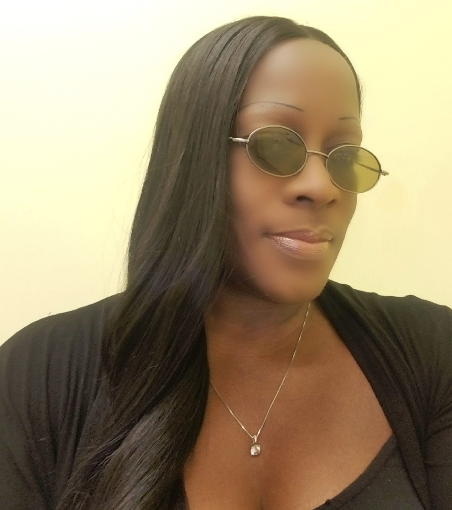 Alyce Barner poses for a close-up photo, wearing circular sunglasses and a necklace, with long, dark hair falling over her shoulder against a pale background.