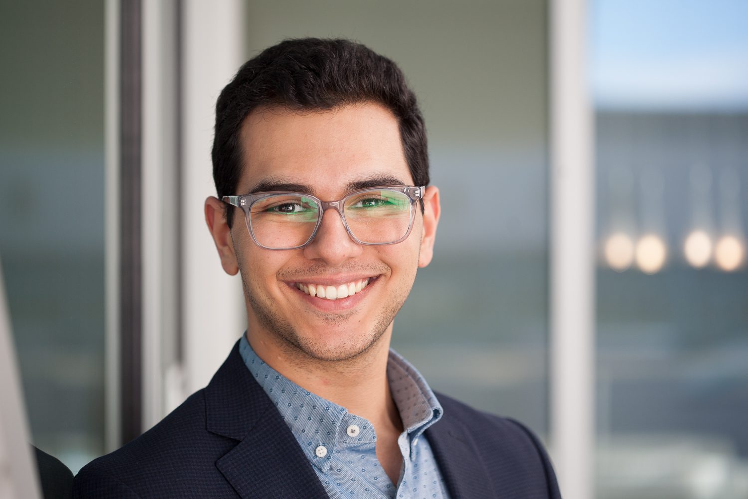 Headshot Arman A. Salehi '23 with glasses smiles while wearing a suit and collared shirt, standing in front of a window with blurred background elements.