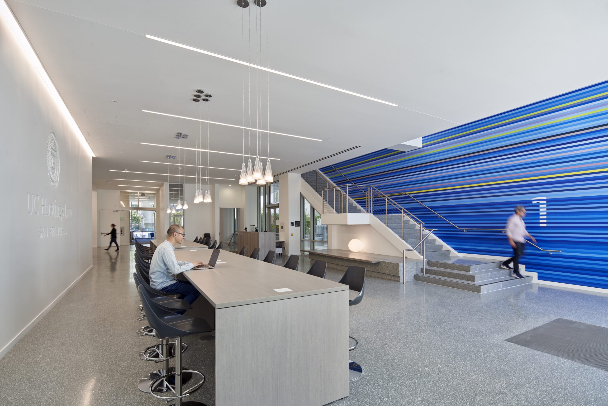 Inside of a white building with a blue wall, made up up stripes in different shades of blue. People are walking and using the tables for studying