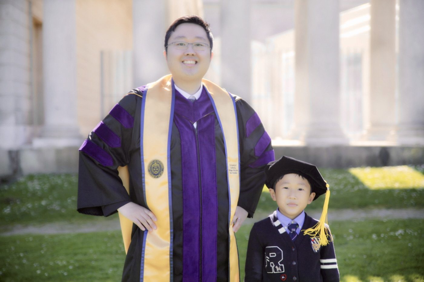 Joseph Jo, '23 wearing a graduation gown and sash, stands next to his five-year-old son, who is dressed in a black suit and cap, outdoors at a historic location.